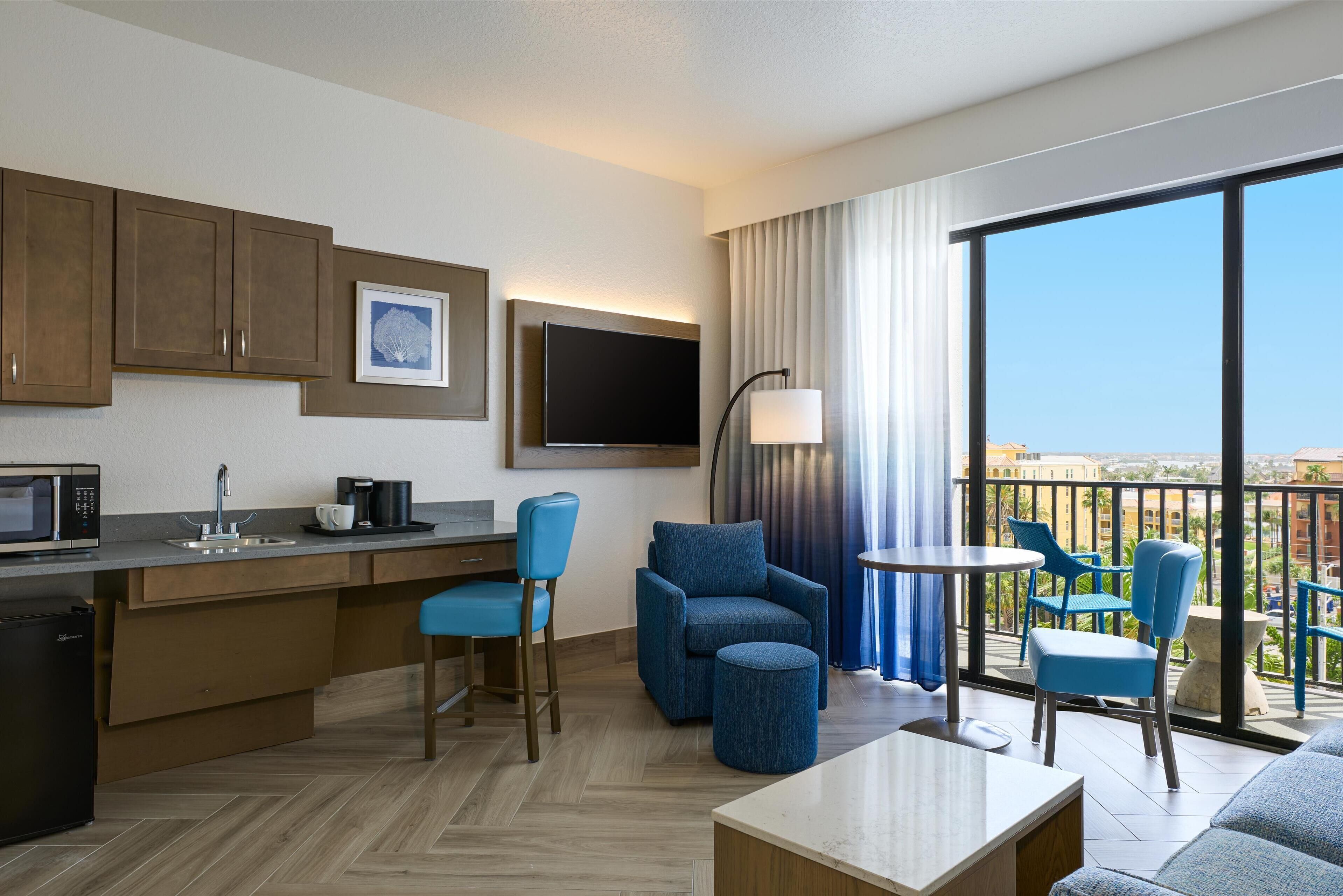 A modern hotel room with a kitchenette, a flat-screen TV, a sofa, a coffee table, and a balcony with a view.
