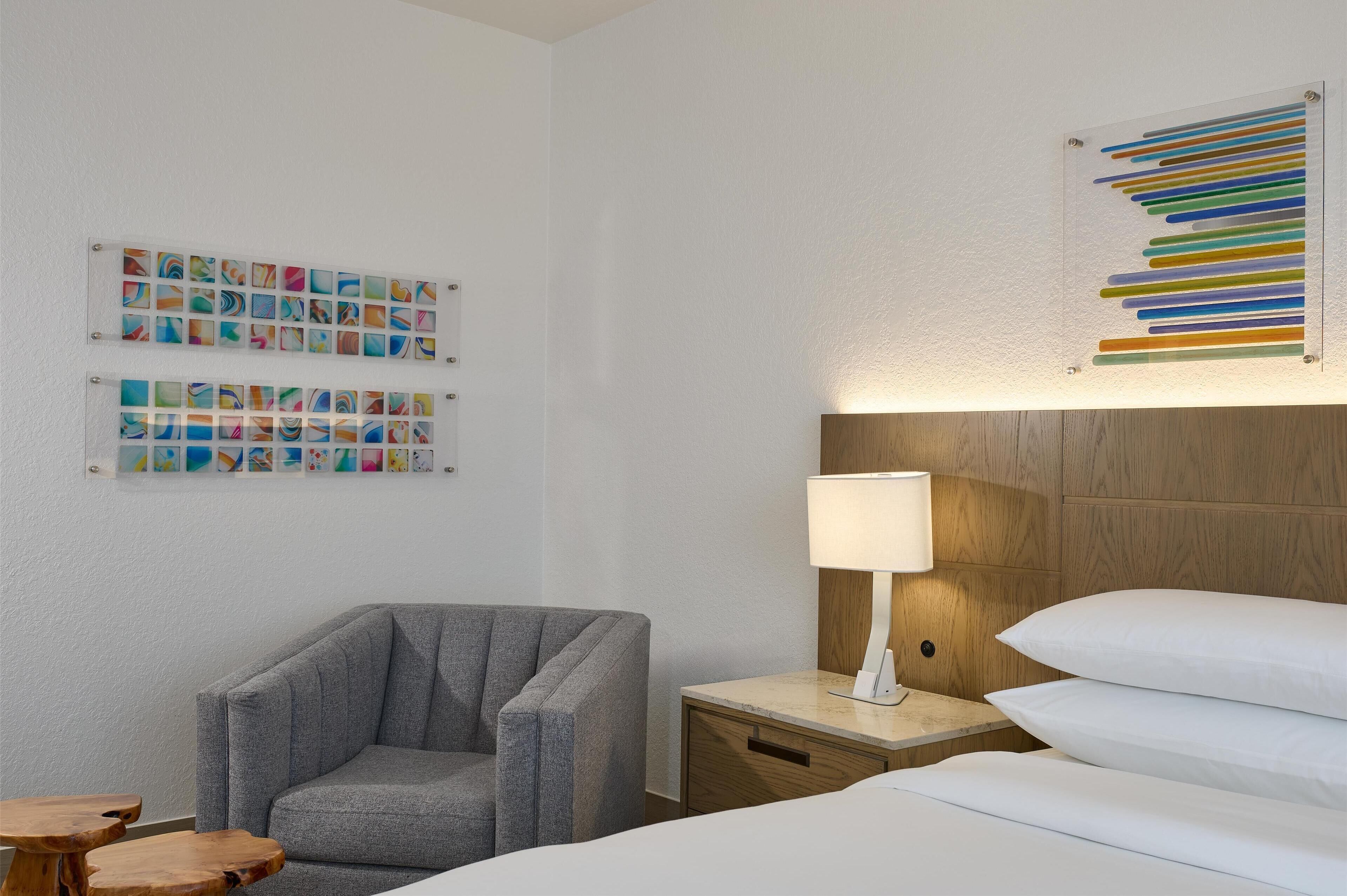 Et hotelværelse med en grå lænestol, et træklædt natbord med en lampe, en seng med hvide sengetøj og to billedrammer på væggen.