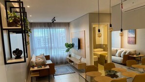 A modern living room with a sofa, coffee table, and dining area. There is a TV mounted on the wall, and the room has a large window with sheer curtains. - Edificio Promenade II - Apto 1101 (Maceió)