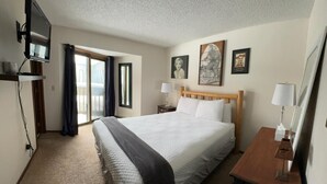 A hotel room with a bed, a TV mounted on the wall, a bedside table with a lamp, and a window with curtains. - Unit 212 - Standard Condo (Winter Park)