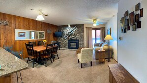 A cozy living room with a fireplace, wooden walls, a dining area with a table and chairs, and a sofa. - Unit 212 - Standard Condo (Winter Park)