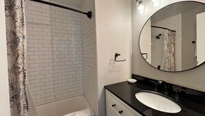Une salle de bain avec une douche carrelée de carreaux blancs, un plan de toilette noir avec un évier et un miroir avec éclairage.