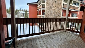 Une terrasse en bois avec une balustrade, un mur en pierre et une vue sur un immeuble à plusieurs étages doté de balcons.