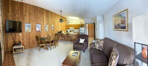 Ein Wohnzimmer mit einer Holzwand, einem Fernseher, einem Essbereich mit Holzmöbeln und einer Küche im Hintergrund.