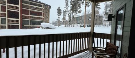 A covered porch with a wooden railing, a bench, and a view of a snow-covered landscape with buildings and trees.