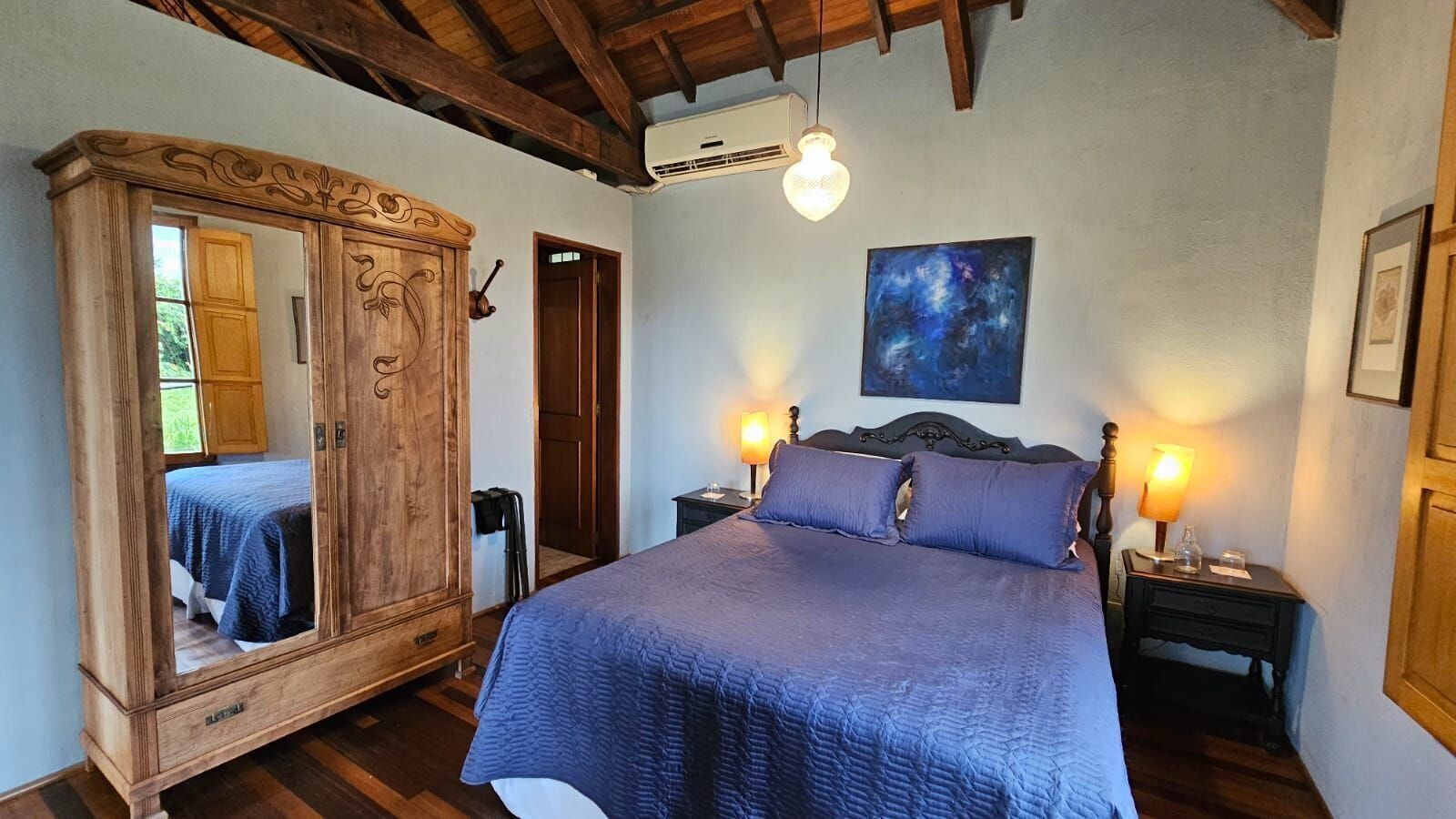 A bedroom with a large wooden wardrobe, a bed with a blue quilt, two bedside tables with lamps, and a painting on the wall.
