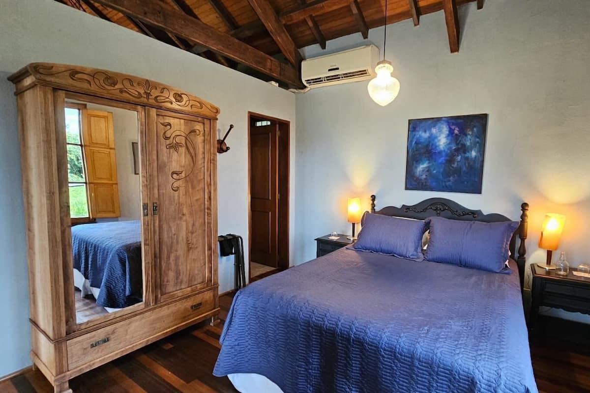 A bedroom with a large wooden wardrobe, a bed with a blue quilt, two bedside tables with lamps, and a painting on the wall.