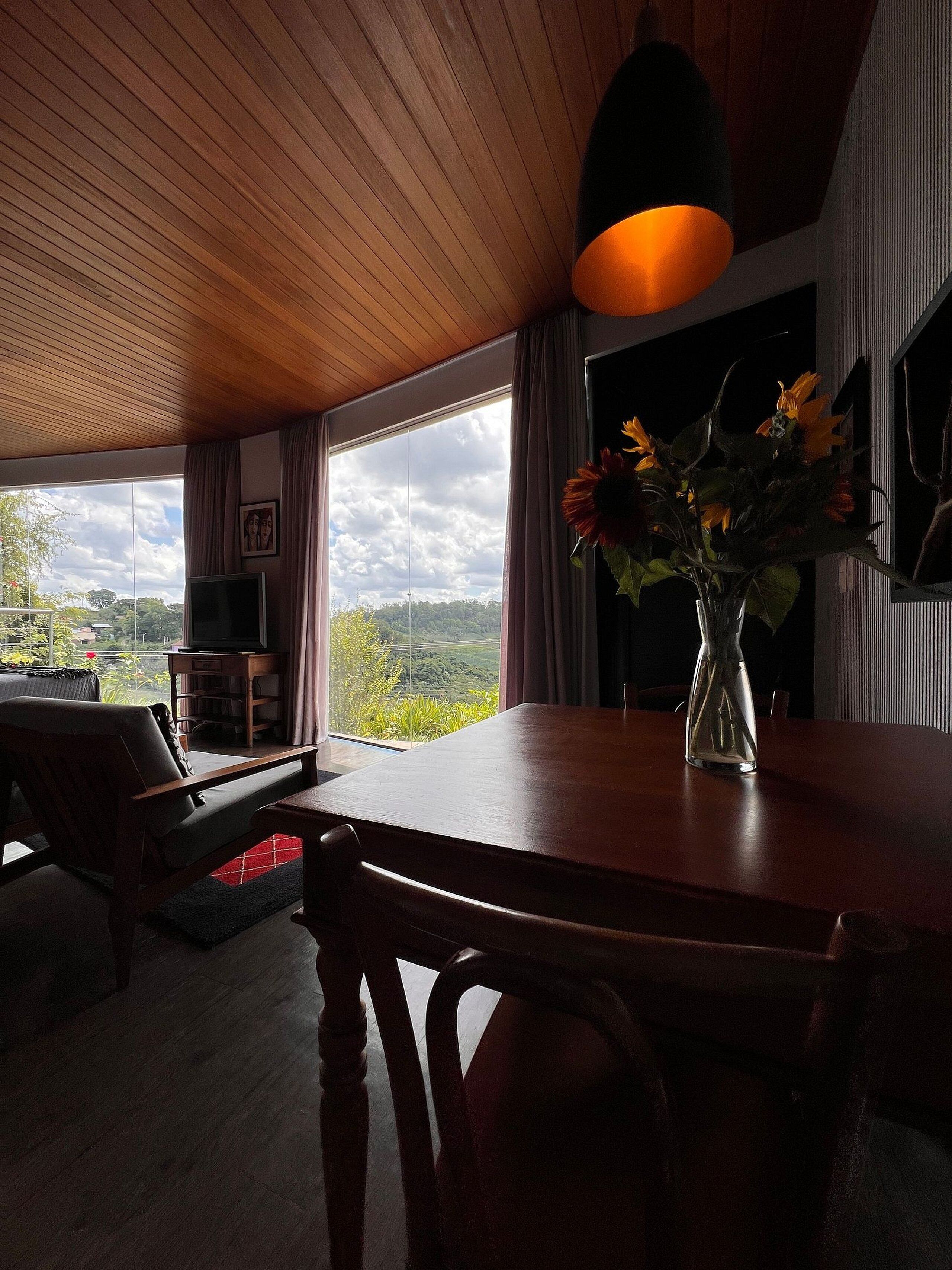 Un comedor con una mesa de madera, sillas, un jarrón con flores, un televisor y vistas al exterior.