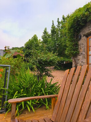 Um deck de madeira com uma cadeira, rodeado por vegetação exuberante e um edifício de pedra.