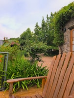 Una terraza de madera con una silla, rodeada de abundante follaje y un edificio de piedra.