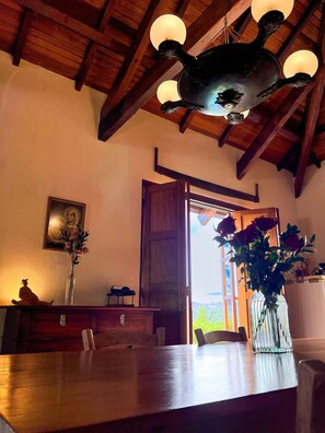 Teto de madeira com um lustre único, mesa de madeira com um vaso de flores e uma porta que dá para uma área externa iluminada.
