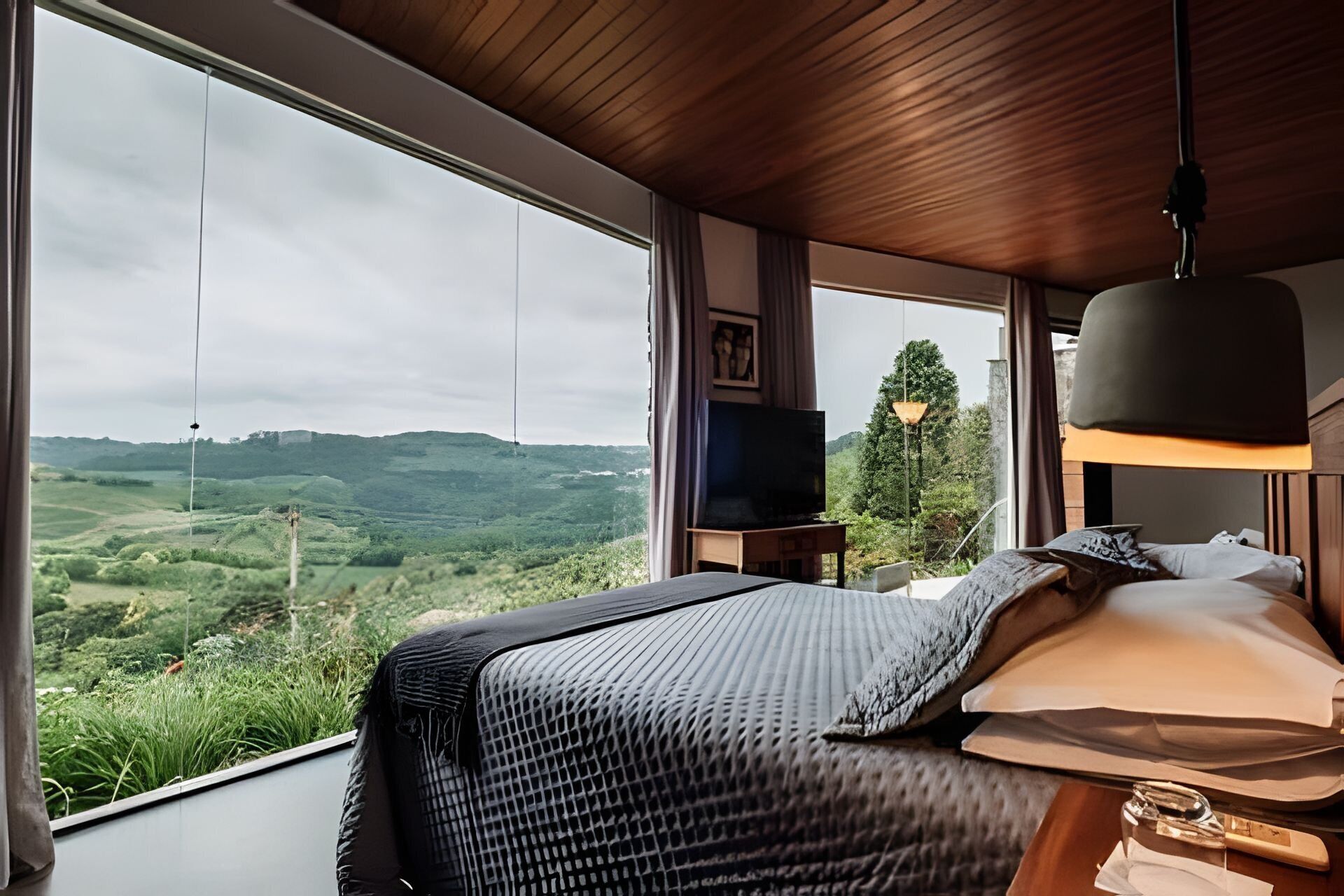 A bedroom with a large window overlooking a green landscape, a bed with a patterned bedspread, a bedside table with a lamp, and a television on a stand.