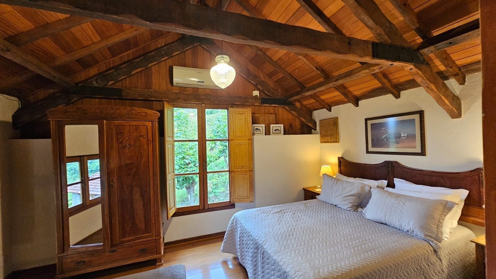 A bedroom with a wooden ceiling, a bed with white bedding, a nightstand with a lamp, and a window with a view of greenery.