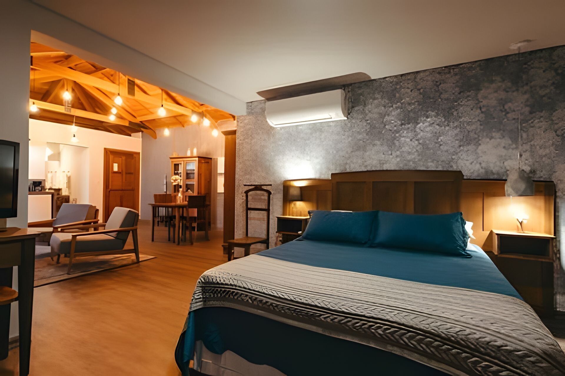 A modern bedroom with a large bed, wooden ceiling beams, a stone accent wall, and a dining area with a table and chairs.