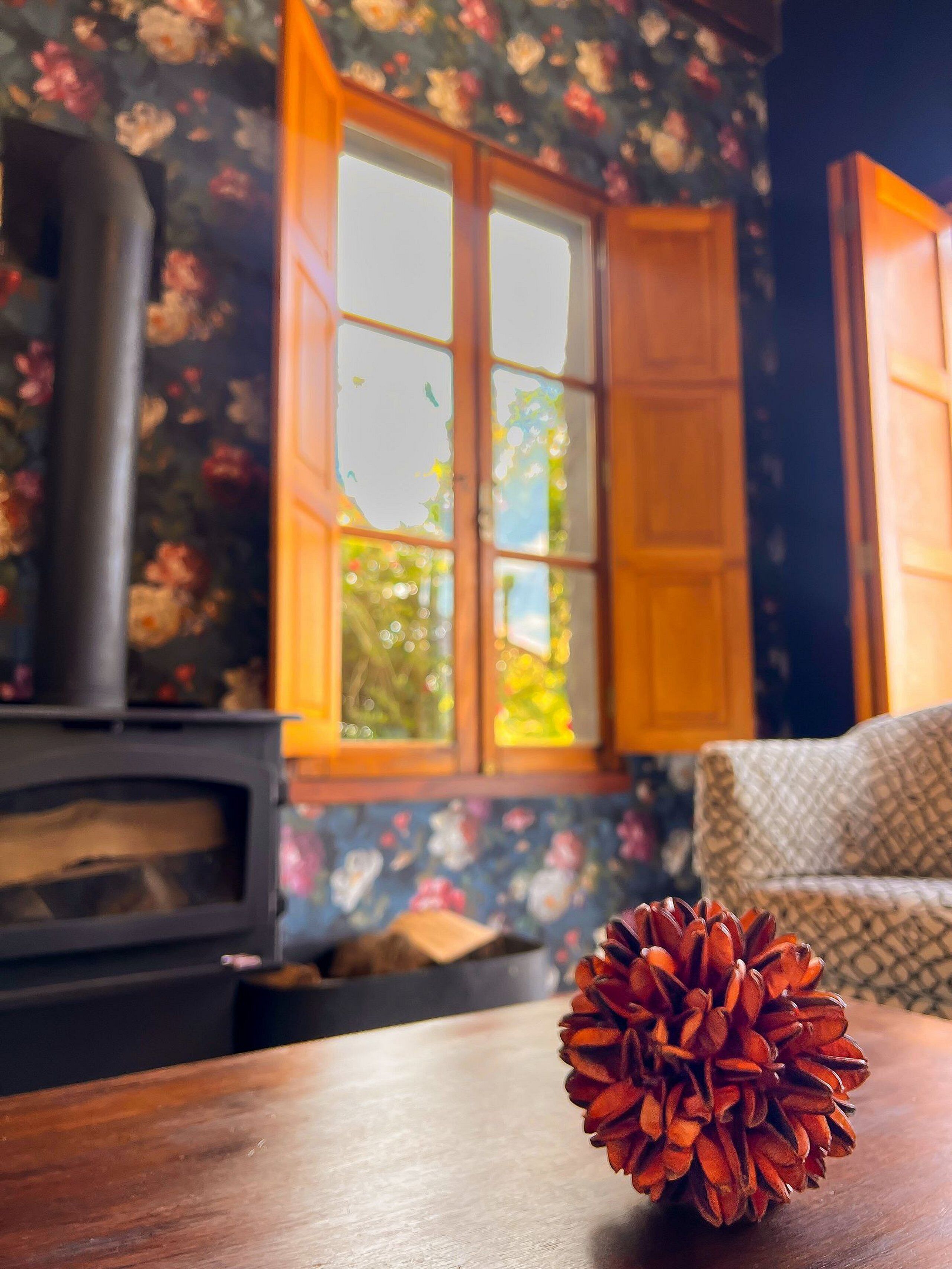 A room with a wooden table, a pine cone, a fireplace, and a window with floral wallpaper.