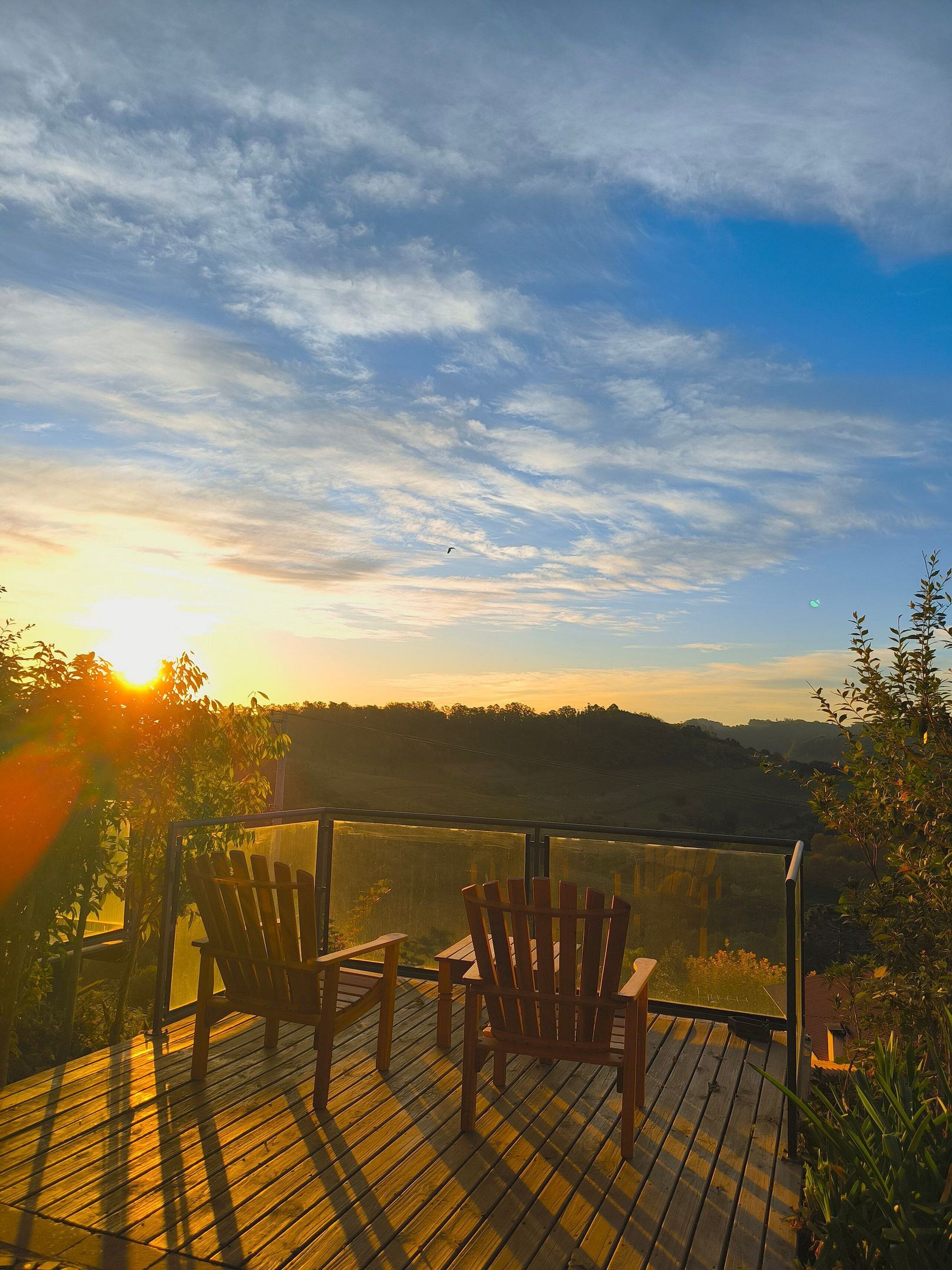 Una terraza de madera con dos sillas orientadas a una vista panorámica de una puesta de sol sobre un paisaje montañoso.