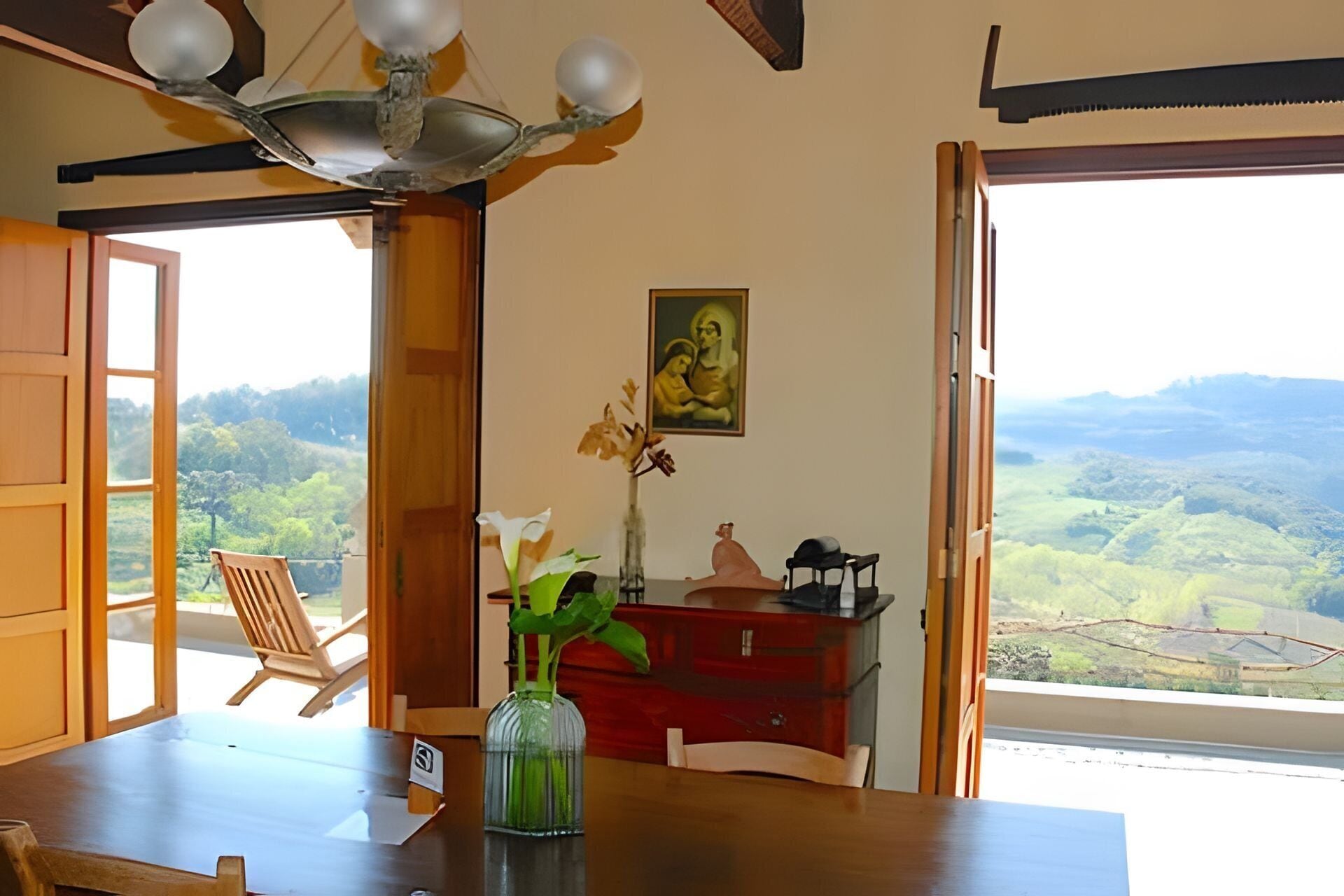 Une pièce avec une table en bois, un vase rempli de fleurs, un buffet rouge et une vue sur un paysage montagneux par une porte ouverte.