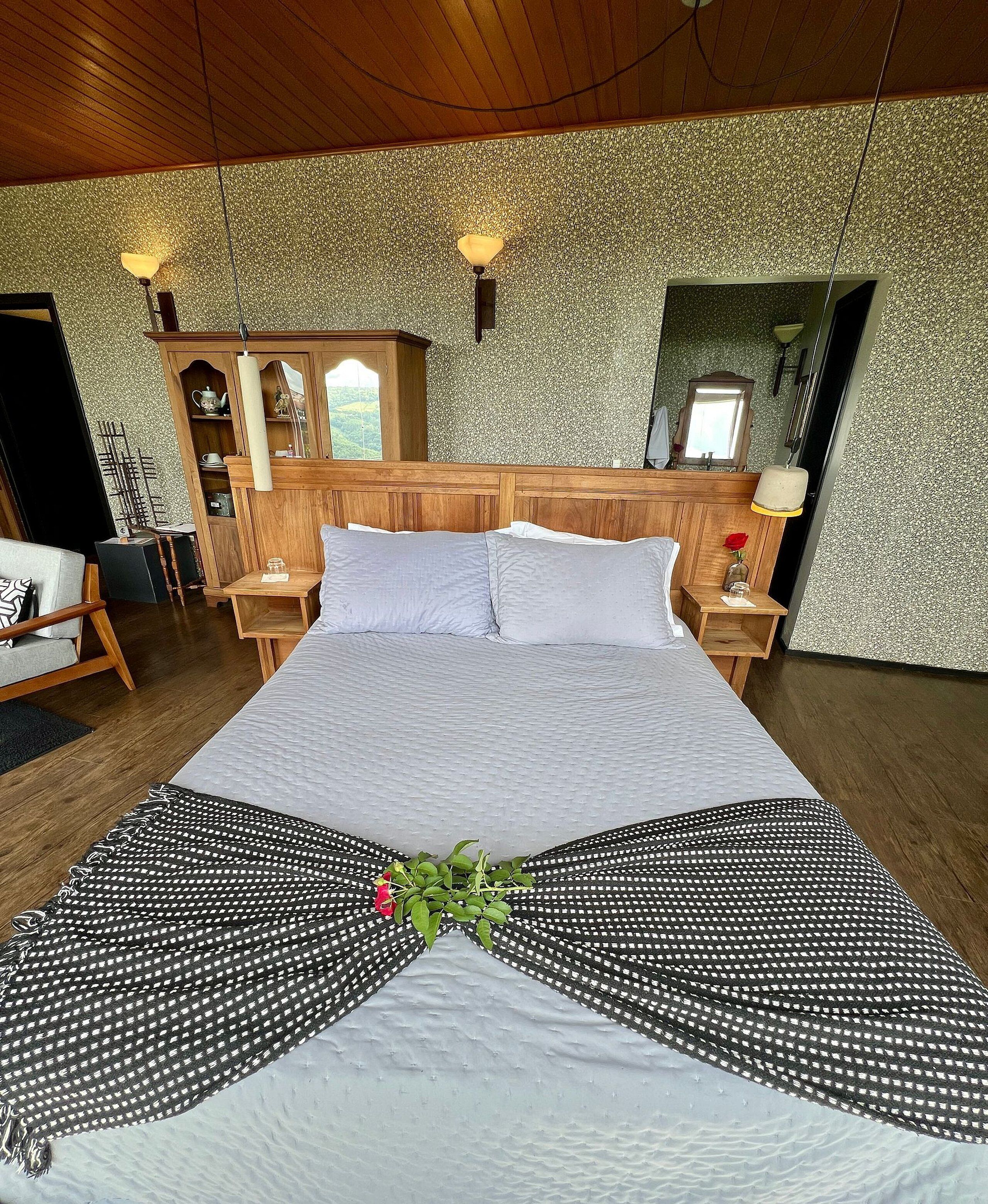 Una cama bien hecha con un arreglo decorativo de almohadas, un cabecero de madera, mesitas de noche con lámparas y vistas al exterior a través de una ventana.