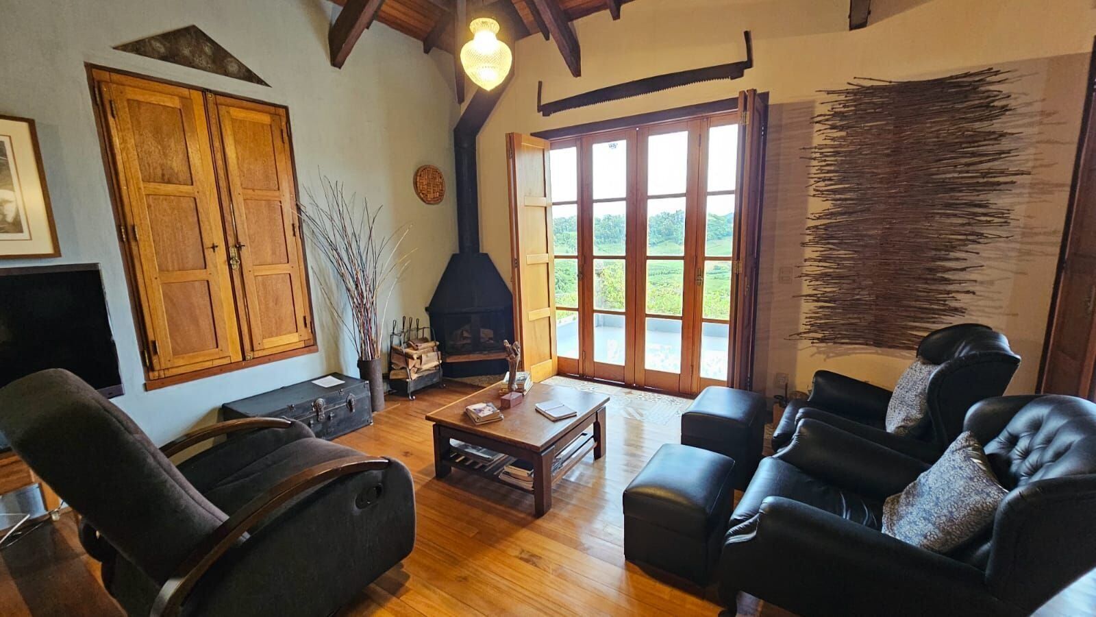 A cozy living room with a fireplace, wooden furniture, and large windows offering a view of the outdoors.