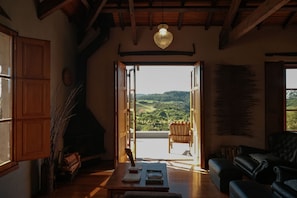 Uma sala de estar acolhedora com teto de madeira, lareira, vista para o campo e um espaço de estar com sofá e poltrona.