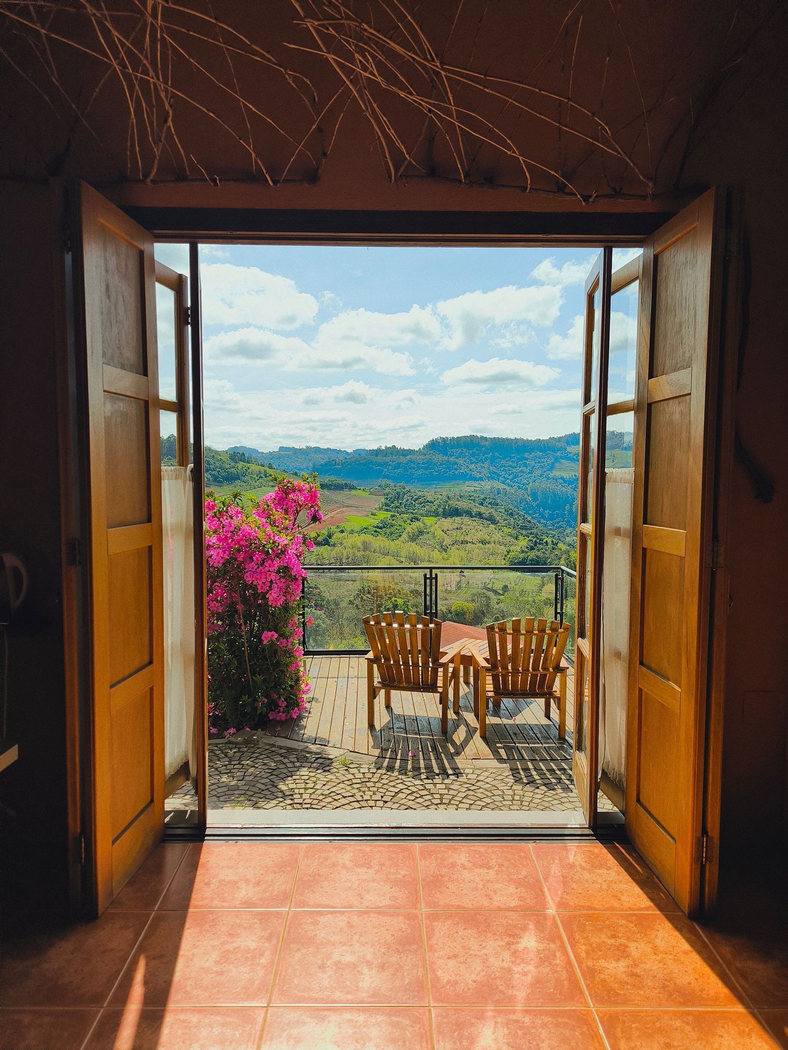 Eine offene Tür, die auf einen Balkon mit Blick auf eine grüne Landschaft und einen blühenden rosa Busch führt.