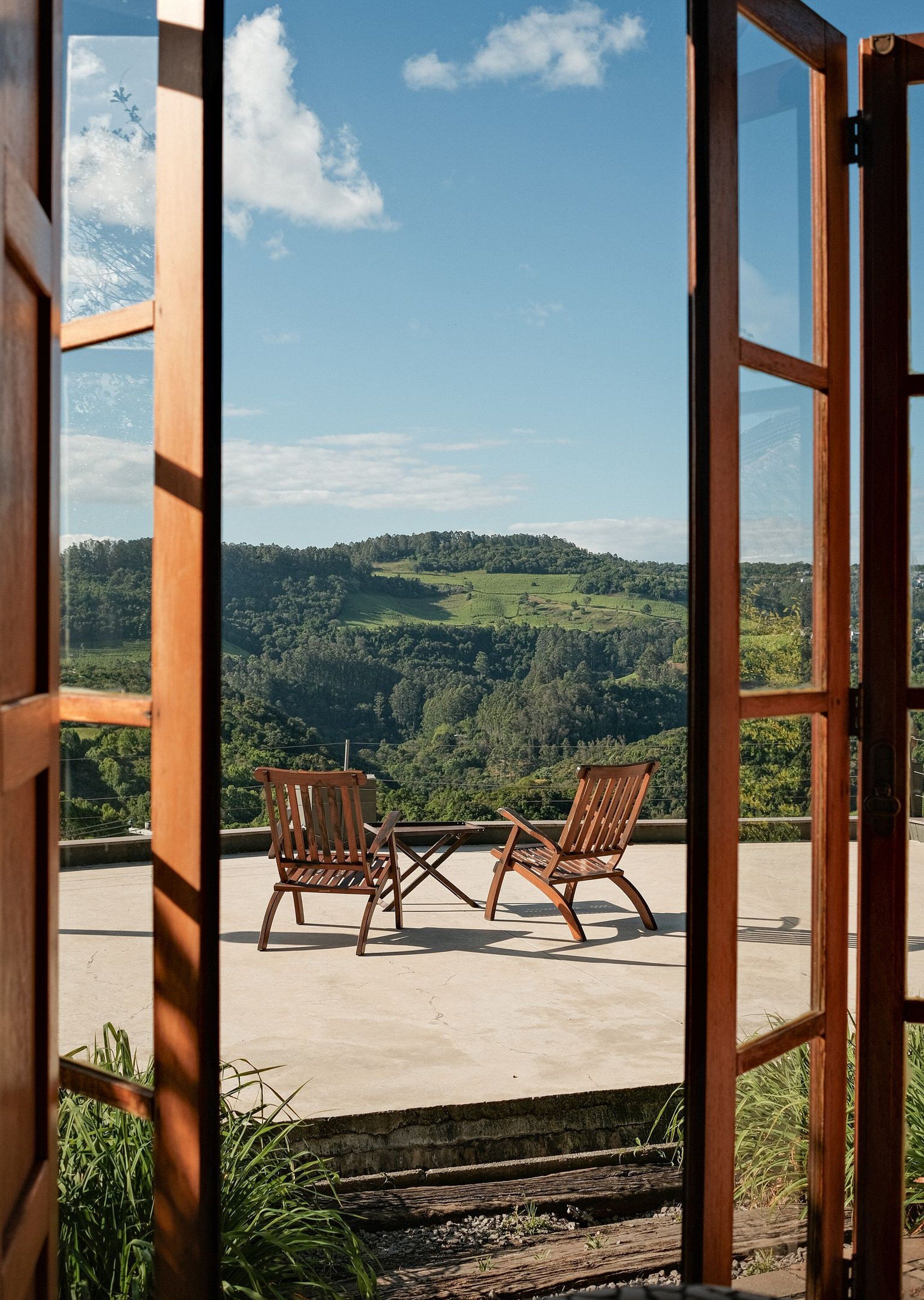Une porte en bois ouverte, avec deux chaises en bois et une petite table sur un balcon donnant sur un paysage verdoyant.