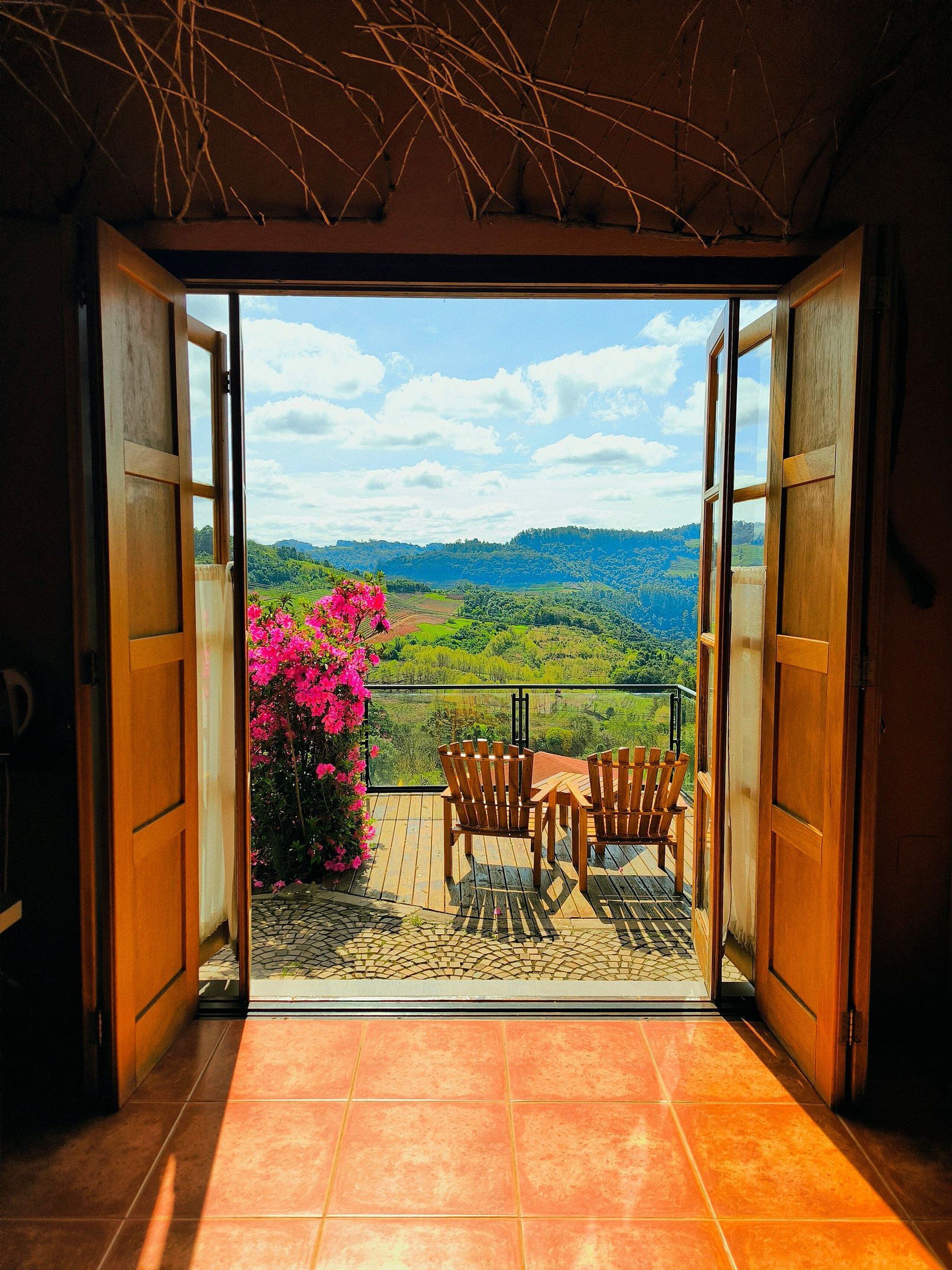 A balcony with a view of a green landscape, two wooden chairs, and a pink flowering plant.