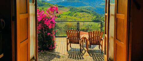 A balcony with a view of a green landscape, two wooden chairs, and a pink flowering plant.