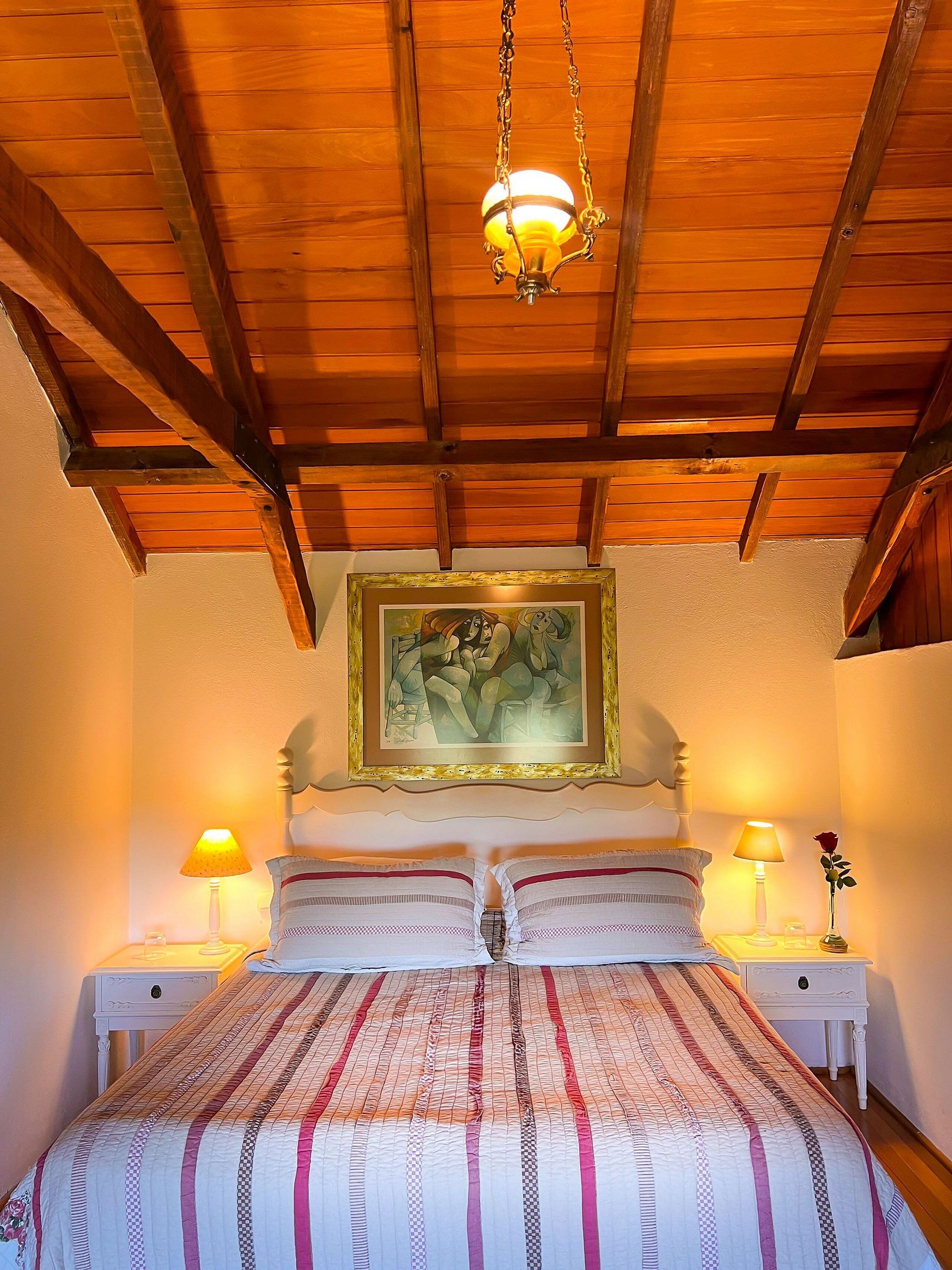 A bedroom with a wooden ceiling, a bed with striped bedding, two bedside tables with lamps, and a framed painting on the wall.