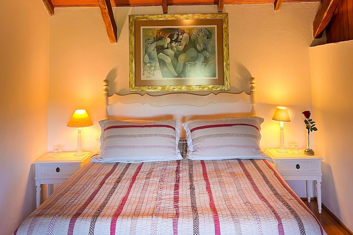 A bedroom with a wooden ceiling, a bed with striped bedding, two bedside tables with lamps, and a framed painting on the wall.