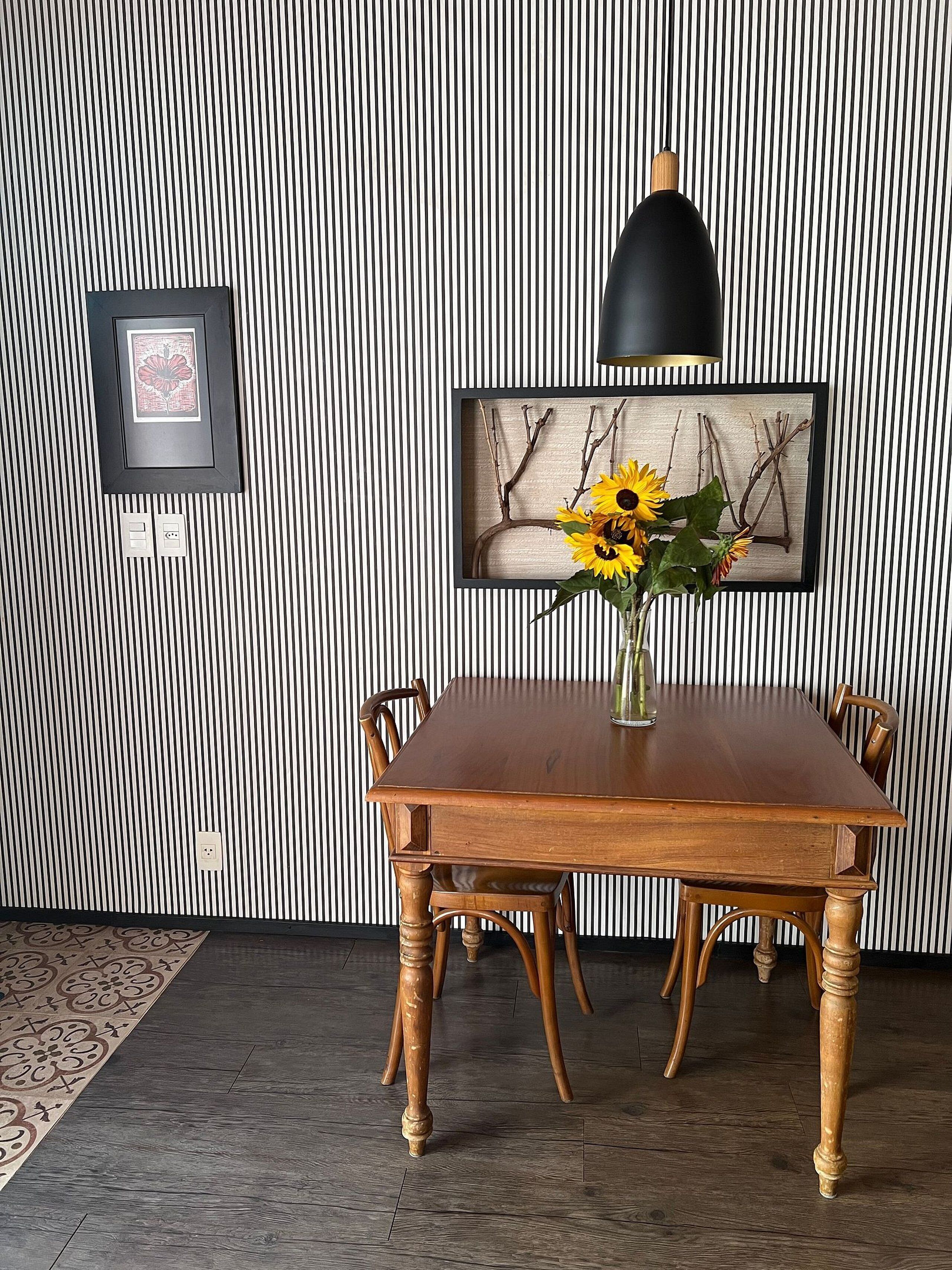 Un comedor con papel tapiz a rayas, un cuadro con ramas, un jarrón con girasoles y una lámpara colgante.