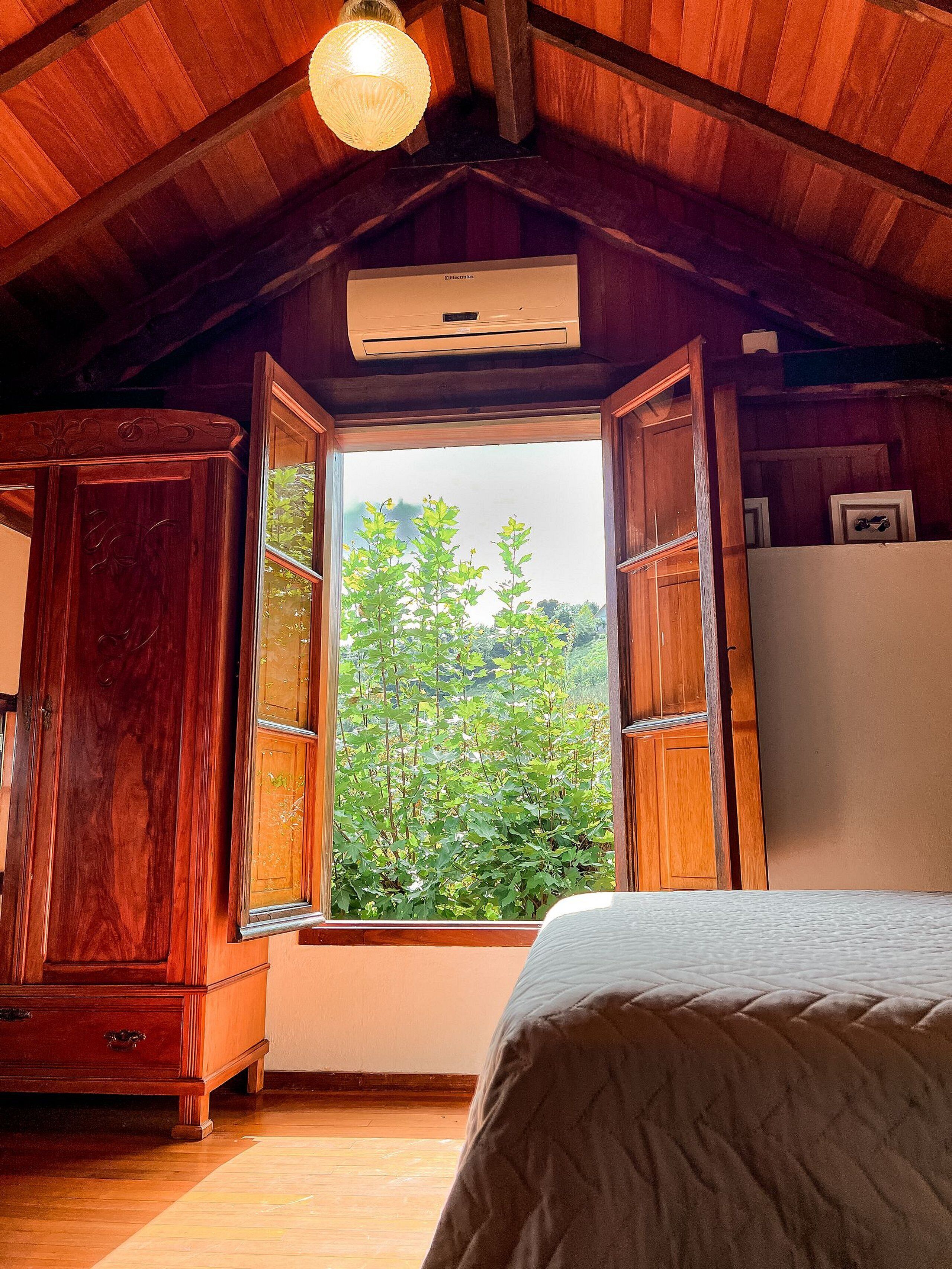 A bedroom with a wooden ceiling, a large window with a view of greenery, a bed, and a wardrobe.