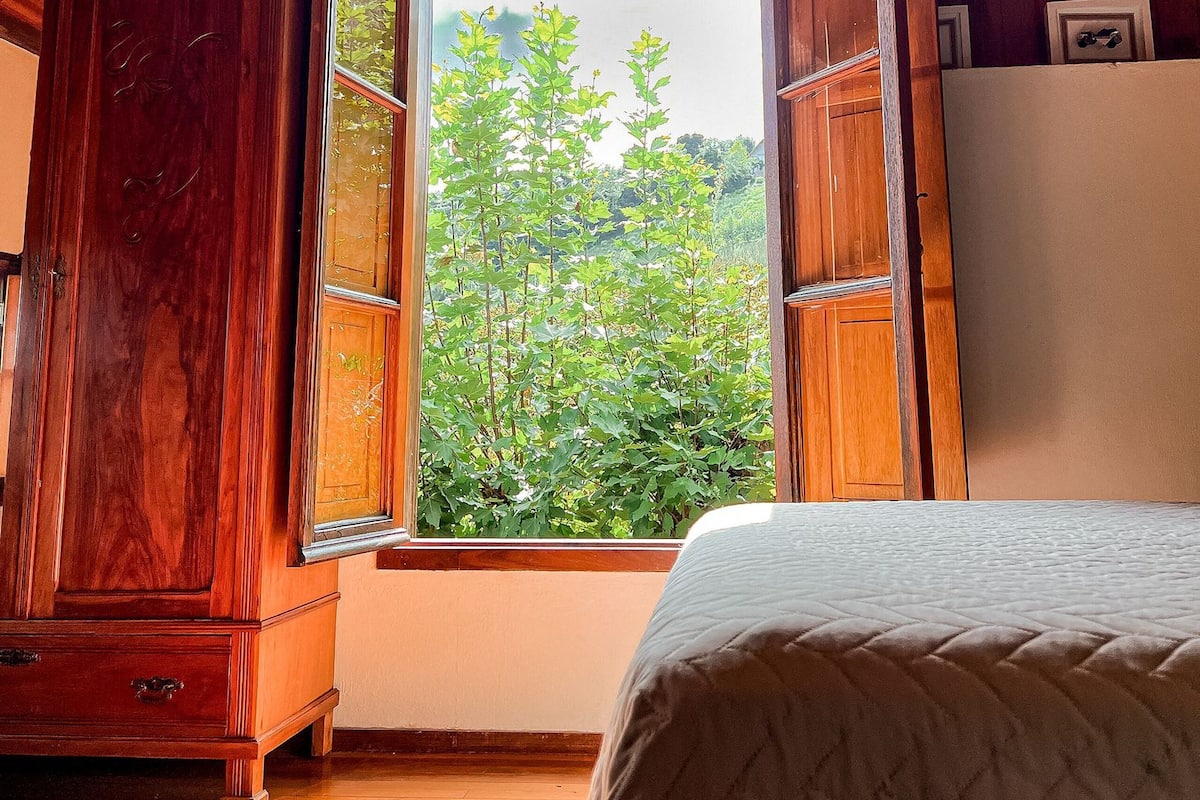 A bedroom with a wooden ceiling, a large window with a view of greenery, a bed, and a wardrobe.