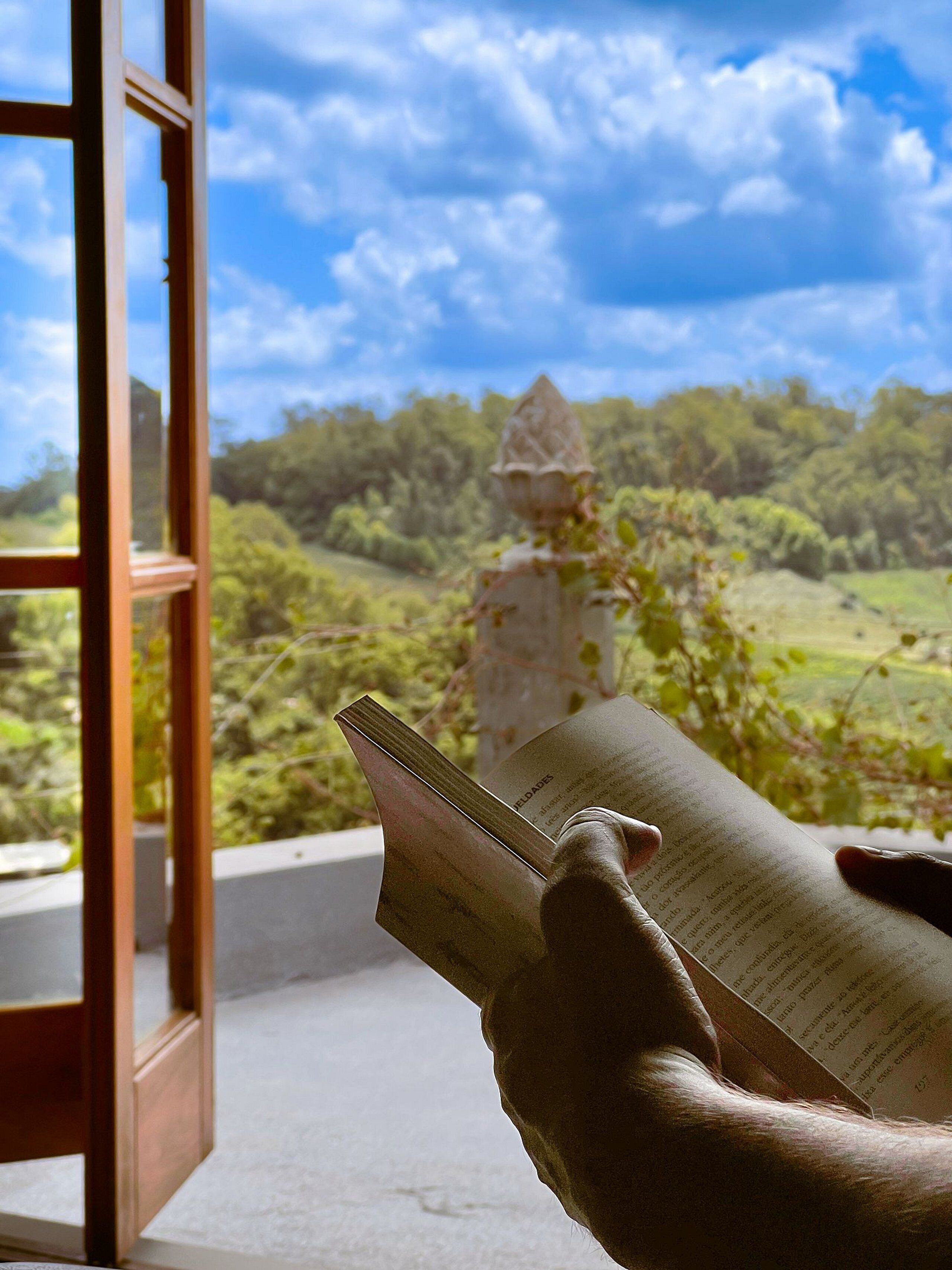 Uma pessoa lendo um livro numa varanda com vista para uma paisagem verde e uma construção de pedra.