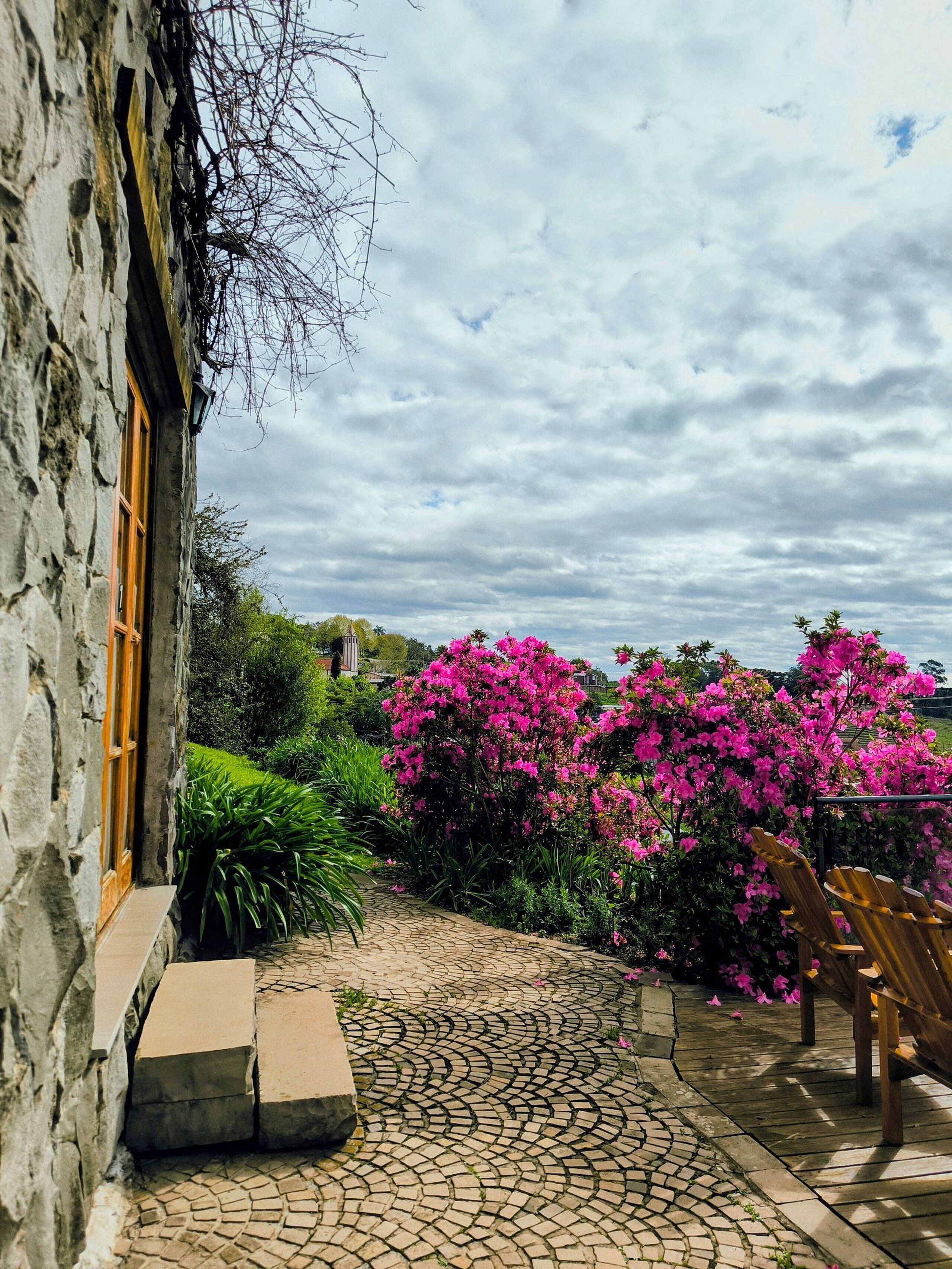 Un edificio de piedra con puerta y ventana de madera, un camino de adoquines y flores rosadas vibrantes al fondo.