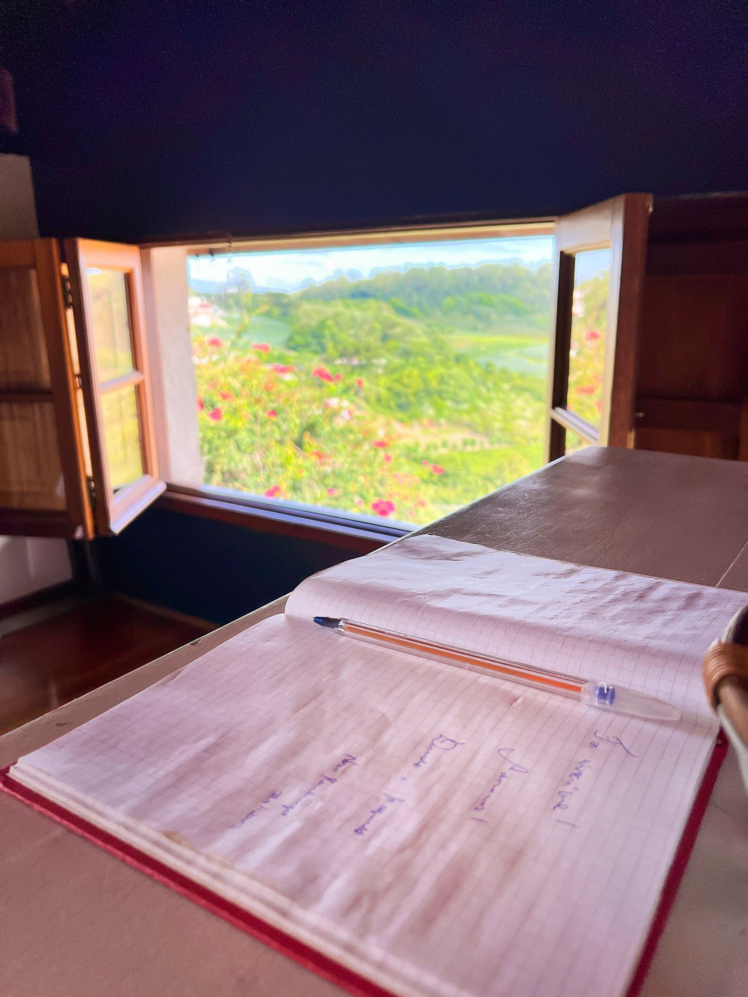 Un cuaderno con un bolígrafo sobre una mesa, una ventana con vistas a zonas verdes, y una puerta de madera.