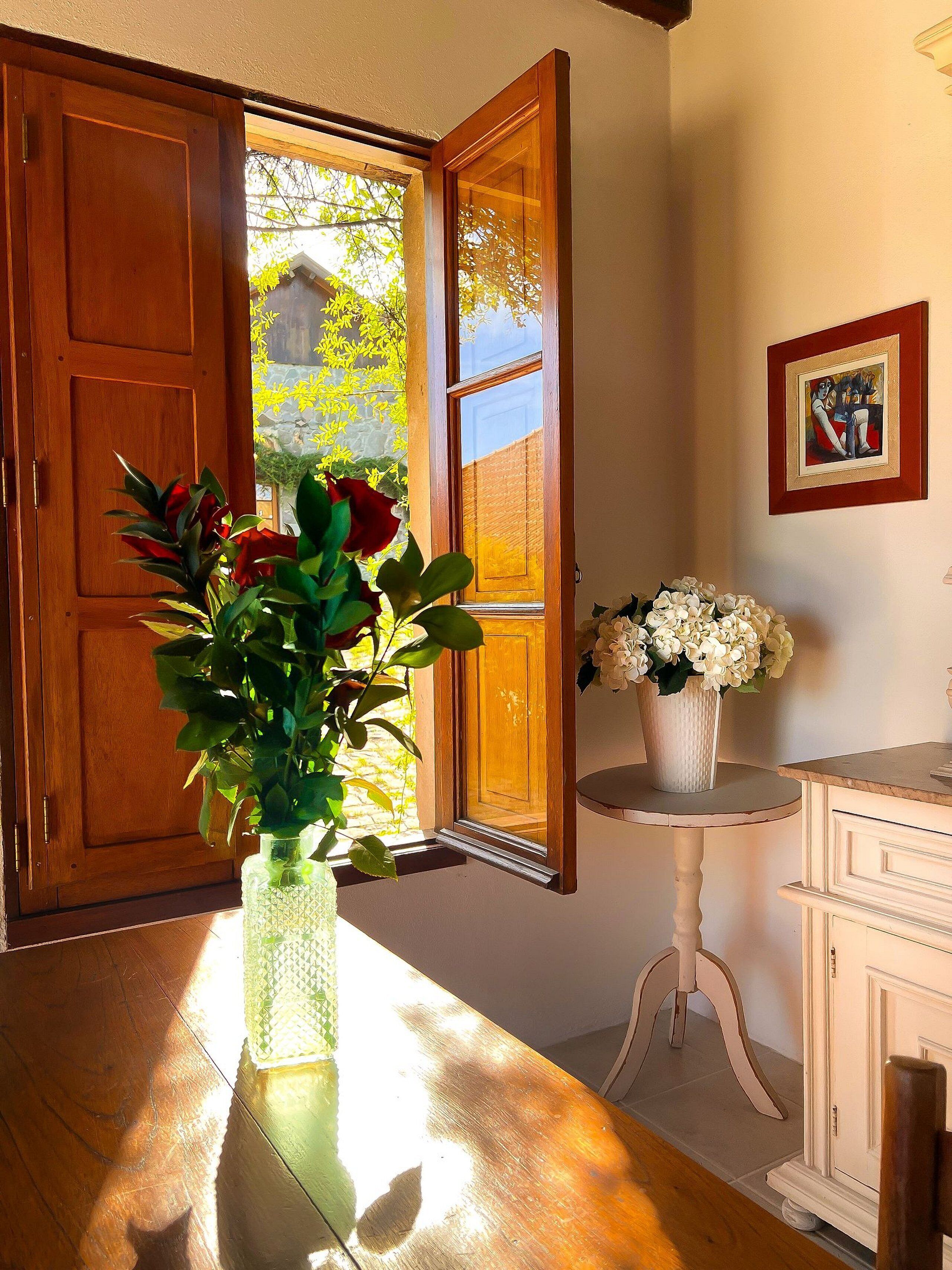 Una puerta de madera abierta que da a un jardín, un florero con flores sobre una mesa y un cuadro en la pared.