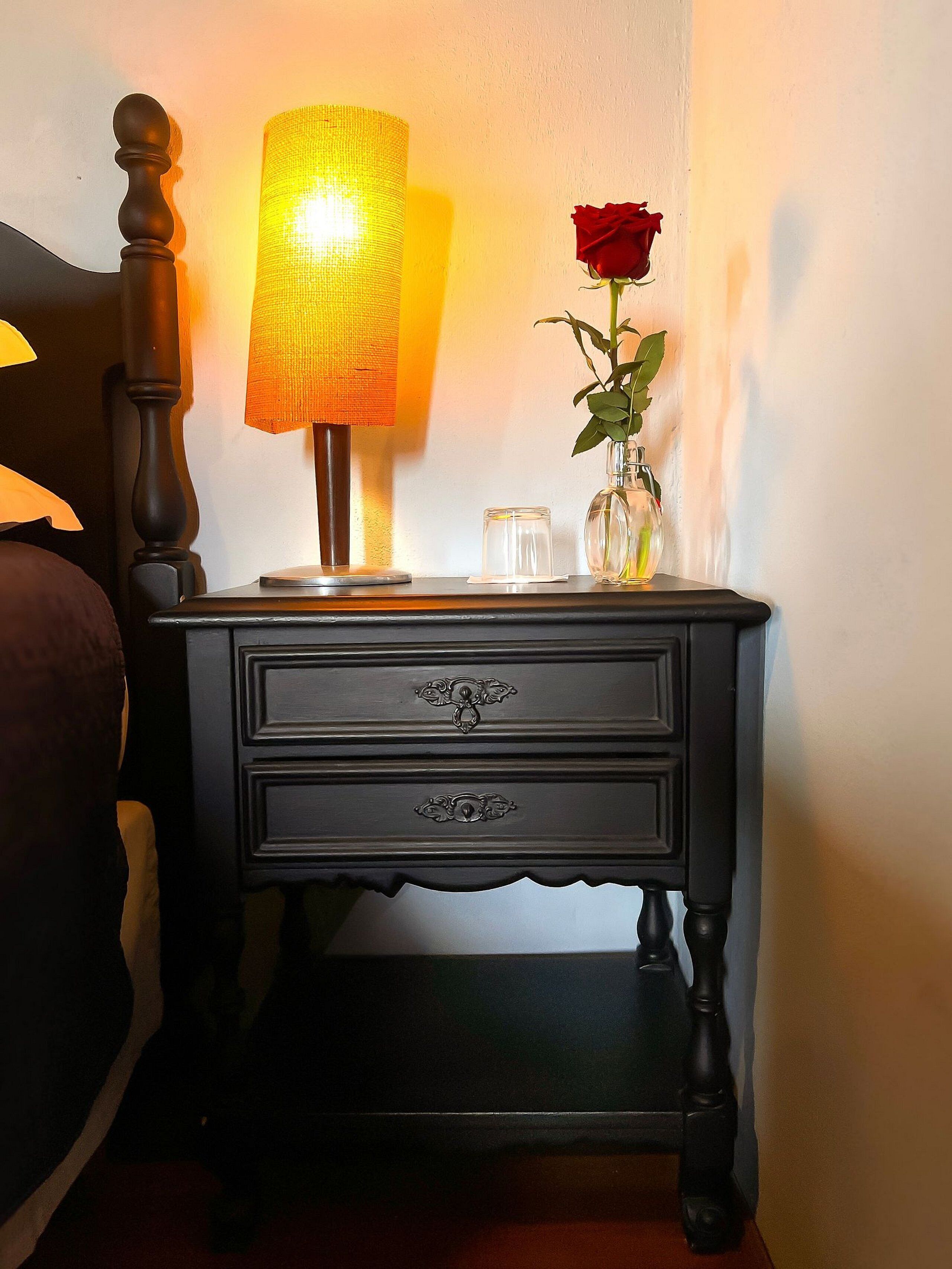 A wooden nightstand with a lamp, a vase with a red rose, and a candle.