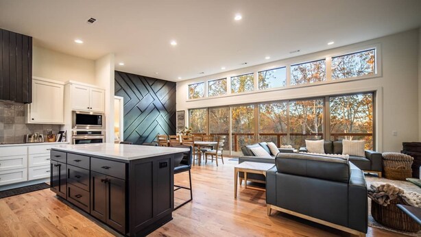 A modern kitchen with a large island, wooden flooring, and a dining area with a view of trees through large windows. - Welcome to PEDAL AND PLUNGE (Bella Vista)