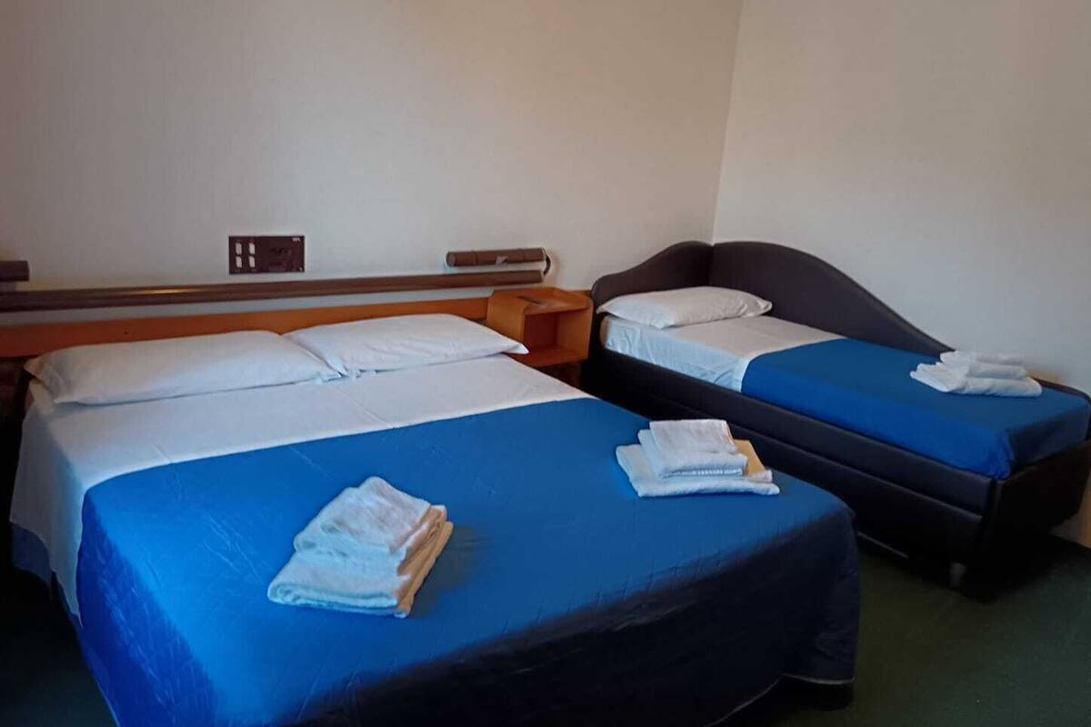 A hotel room with two single beds, each with a blue bedspread and white pillows. There are folded towels on the beds.