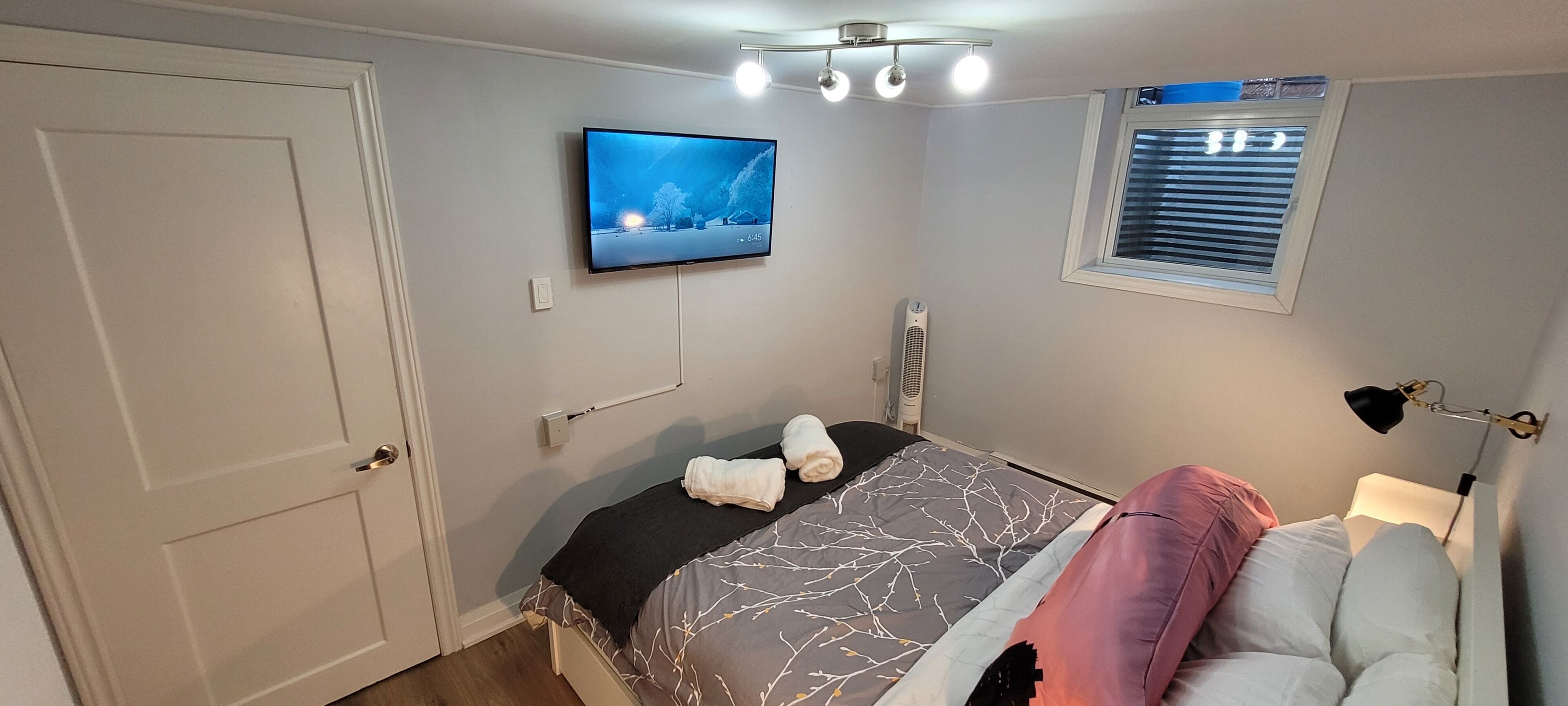 A bedroom with a bed, a TV mounted on the wall, a window with blinds, and a ceiling light fixture.