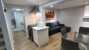 A compact kitchen with a microwave, stove, and a small refrigerator. There is a dark-colored sofa and a glass dining table with chairs. A painting is visible on the wall. - A Basement Studio for 2 (Brampton)