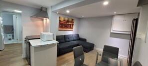 Un espace de vie compact comprenant un coin cuisine, un canapé noir, une table à manger en verre et un tableau accroché au mur.