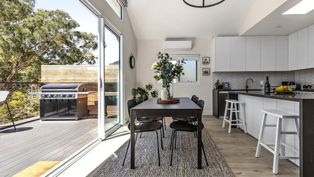 Une cuisine moderne avec des meubles blancs, un coin repas avec une table et des chaises, un barbecue à l’extérieur et une vue sur les arbres.