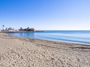 A sandy beach with footprints leading to a rocky breakwater with buildings and palm trees. - Cubo's Beach Side Trinidad Apartment / close to the beach / free wifi (Fuengirola)