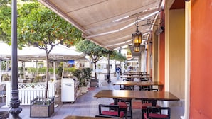 Un café al aire libre con mesas y sillas de madera, una zona cubierta para sentarse y vistas a la calle con tiendas y árboles.