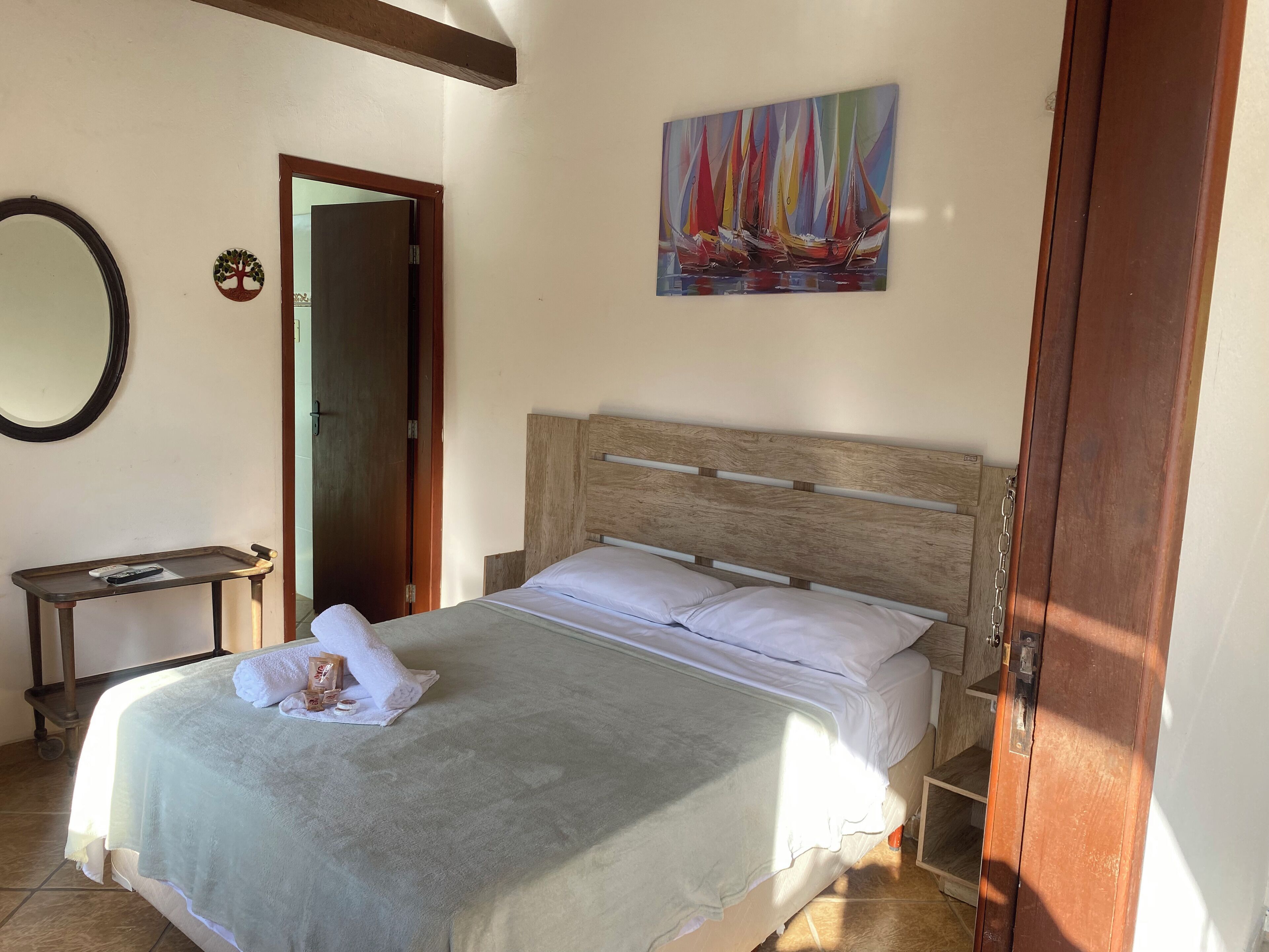 A neatly made bed with a wooden headboard, a bedside table with a remote control, and a painting of sailboats on the wall.