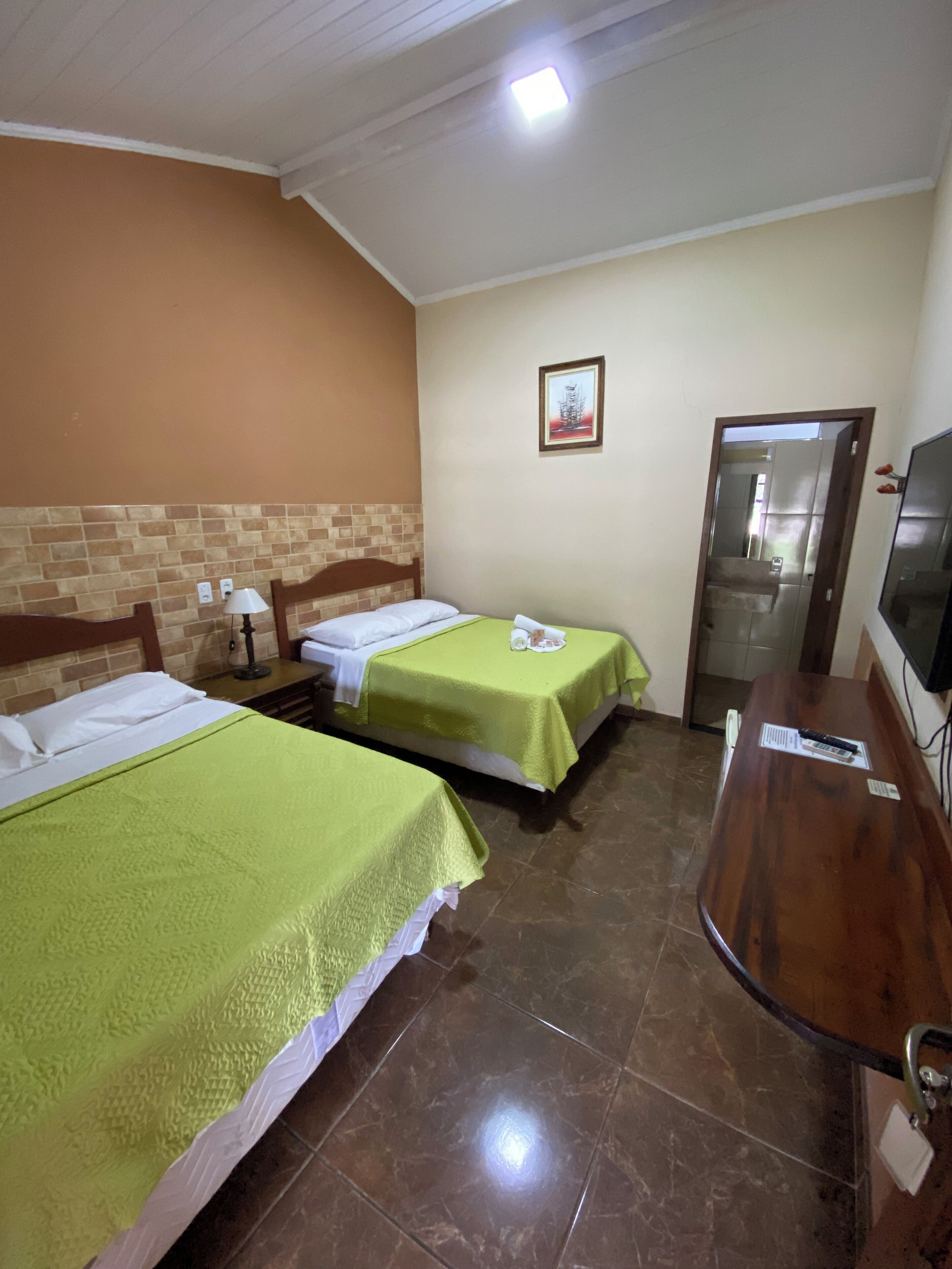 A hotel room with two beds, a wooden headboard, a small table with a lamp, a television, and a door leading to a bathroom.