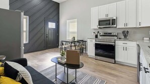 Una cocina moderna con armarios blancos, un microondas y un horno. Cuenta con una mesa de comedor con sillas y un sofá negro con cojines amarillos.