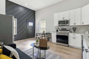 A modern kitchen with white cabinets, a microwave, and an oven. There is a dining table with chairs and a black sofa with yellow cushions.